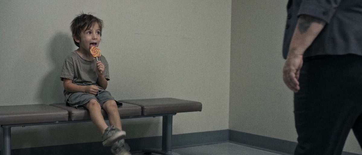 A child eating a lollipop on a waiting-room bench as an adult stands nearby