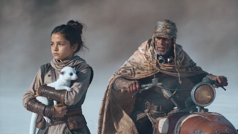 Kira holds a small white creature beside a cloaked rider and motorcycle